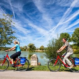 Zwei Radfahrer auf roten Fahrrädern mit blauen Taschen fahren auf einem asphaltierten Weg entlang eines Sees in Bayern, mit Bäumen und blauem Himmel im Hintergrund.