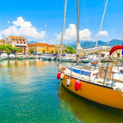 Gelbes Segelboot mit roten Fendern im Hafen von Saint-Florent, Korsika. Bunte Gebäude säumen den Hafen mit Bergen im Hintergrund.