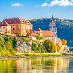 Dürnstein auf Felsenklippe über der Donau mit barockem blauen Kirchturm und historischen Gebäuden, grüne Hügel im Hintergrund.
