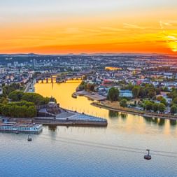Luftaufnahme des Deutschen Ecks in Koblenz bei Sonnenuntergang mit Flusskonvergenz, Seilbahnen, Kreuzfahrtschiffen und der Stadt unter goldenem Himmel.