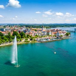 Luftaufnahme von Friedrichshafen am Bodensee mit Springbrunnen, historischen Gebäuden, Kirchturm und Fahrgastschiff auf türkisem Wasser.