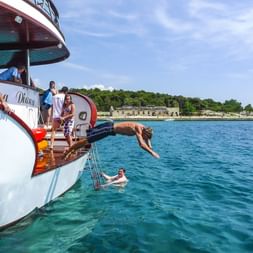 Person springt von einem Boot ins türkisfarbene Wasser der Kvarner Bucht, Kroatien. Andere Personen an Deck und schwimmend, Küstenlandschaft sichtbar.
