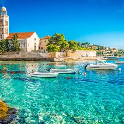 Franziskanerkloster in Hvar mit Glockenturm an der Uferpromenade. Kristallklares türkisfarbenes Wasser mit vertäuten Booten im Vordergrund.