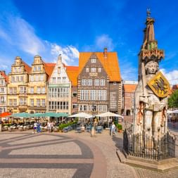 Bremer Marktplatz mit historischer Rolandstatue im Vordergrund, bunten Giebelhäusern, Straßencafés mit orangen Schirmen und blauem Himmel.
