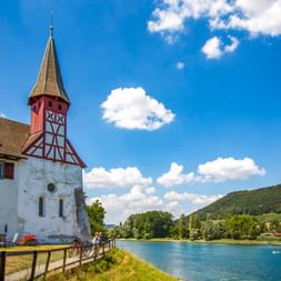 Radfahrer auf einem Uferweg neben einer weißen Kirche mit rot-weißem Fachwerk-Turm in Stein am Rhein. Der Rhein und grüne Hügel unter blauem Himmel.