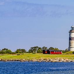 Morups Tånge Leuchtturm mit weißen und braunen Streifen auf grüner Küstenlandschaft. Rote Gebäude und felsige Küste mit blauem Wasser.