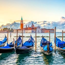 Row of blue gondolas moored at wooden poles in Venice with San Giorgio Maggiore church across the lagoon under a dramatic sunset sky.