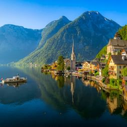 Hallstatt mit Kirchturm am Hallstätter See, umgeben von bewaldeten Bergen. Ein kleines Boot fährt über das ruhige Wasser, das die Szenerie spiegelt.