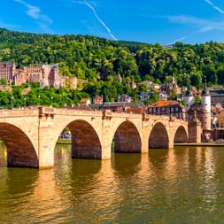 Historische Steinbogenbrücke über den Neckar in Heidelberg mit Schlossruine am bewaldeten Hang und Kirchturm in der Altstadt.