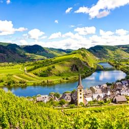 Dorf Bremm mit Kirchturm in der Moselschleife, umgeben von terrassierten Weinbergen und grünen Hügeln unter blauem Himmel.