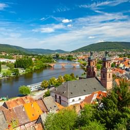 Luftaufnahme von Miltenberg mit historischen Gebäuden, roten Dächern und Kirchtürmen am Main, umgeben von grünen Hügeln.