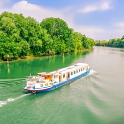Weiß-blaues Passagierschiff L'Estello auf einem grünen Fluss in Frankreich, umgeben von üppigen Bäumen unter blauem Himmel mit weißen Wolken.