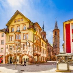 Stadtplatz in Villingen-Schwenningen mit historischem gelbem Gebäude, Münsterturm und modernem rot-weißem Gebäude unter blauem Himmel.