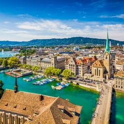Luftaufnahme von Zürich mit der türkisfarbenen Limmat, die durch die Stadt fließt, historische Kirche mit grünem Turm, Boote am Fluss.