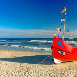 Rotes Fischerboot mit bunten Flaggen am Sandstrand in Ahlbeck, Usedom. Blaue Ostsee mit sanften Wellen unter klarem Himmel.