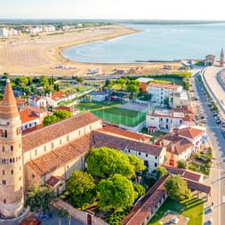 Luftaufnahme von Caorle mit historischem Glockenturm, Küstenpromenade, Sandstrand mit Sonnenschirmen und der Adria.