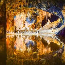 Beleuchtetes Inneres der Saalfelder Feengrotten mit bunten Stalaktiten und Stalagmiten, die sich in stillem unterirdischem Wasser spiegeln.