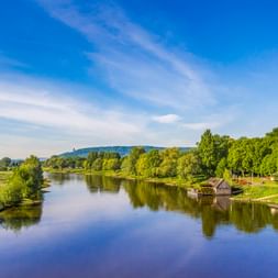 Ruhige Weser mit historischer Schiffmühle am rechten Ufer, grünen Wiesen, Bäumen und fernen Hügeln unter blauem Himmel bei Minden.