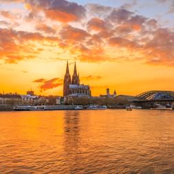 Kölner Skyline im Abendlicht mit Dom, Groß St. Martin und Hohenzollernbrücke, die sich im Rhein unter dramatischen orangefarbenen Wolken spiegeln.