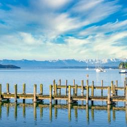Holzstege ragen in den Starnberger See mit Segelbooten im ruhigen Wasser. Schneebedeckte Alpen am Horizont unter blauem Himmel.