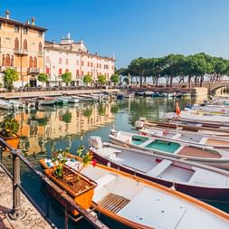Hafen in Desenzano del Garda mit vertäuten Booten, bunten italienischen Gebäuden und Bäumen entlang der Uferpromenade unter blauem Himmel.