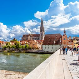 Fußgänger und Radfahrer auf der Steinernen Brücke über die Donau in Regensburg mit historischem Dom und bunten Gebäuden.