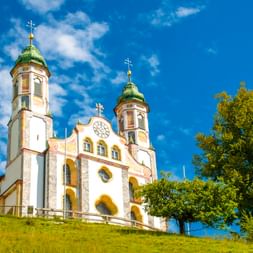 Weiße Barockkirche mit zwei grünen Kuppeltürmen auf einem Grashügel bei Bad Tölz in Oberbayern, umgeben von Bäumen unter blauem Himmel.