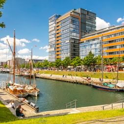 Historische Segelschiffe am Kieler Germaniahafen mit modernen Gebäuden und grüner Uferpromenade unter blauem Himmel mit weißen Wolken.