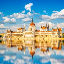 Ungarisches Parlamentsgebäude in Budapest mit weißer neugotischer Fassade und orangen Kuppeln, gespiegelt in der ruhigen Donau unter blauem Himmel.