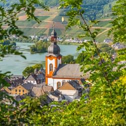 Blick auf Zell an der Mosel durch grünes Laub, mit Kirche mit orangefarbenem Turm und Kuppel an der Mosel mit Weinberghügeln.