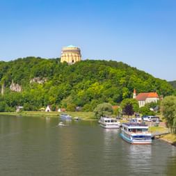Befreiungshalle auf bewaldetem Hügel über Kelheim mit Donau, anlegenden Kreuzfahrtschiffen und Kirche im Uferort unter blauem Himmel.