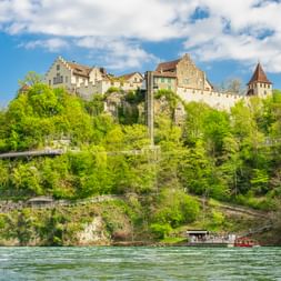 Schloss Laufen thront auf einer Felsklippe über dem Rhein bei Neuhausen am Rheinfall, umgeben von üppig grünen Bäumen unter blauem Himmel.