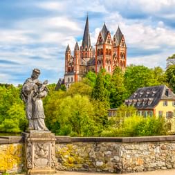 Steinstatue auf historischer Brücke mit Blick auf den Limburger Dom mit sieben Türmen. Gelbes Wohnhaus und grüne Bäume rahmen die rote Sandsteinkirche.