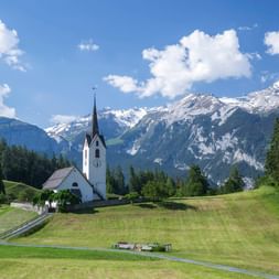 Weiße Kirche mit hohem Turm in Versam, umgeben von grünen Wiesen und Wäldern, mit schneebedeckten Schweizer Alpen unter blauem Himmel.