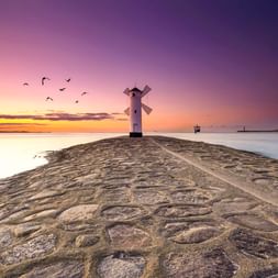 Mühlenförmige Bake auf Steinmole in Swinemünde bei Sonnenuntergang. Vögel fliegen über lila-orangefarbenem Himmel über ruhiger Ostsee.