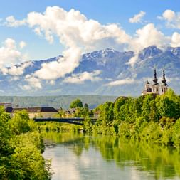 Blick auf Villach mit grünem Fluss, üppigen Bäumen, Kirche mit Zwillingstürmen und schneebedeckten Bergen unter bewölktem Himmel.