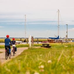 Zwei Radfahrer mit roten Helmen fahren auf einem Küstenweg in Nordstrand, vorbei an einem Yachthafen mit Segelbooten und grünen Wiesen.