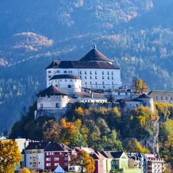 Festung Kufstein auf einem Felsenhügel über bunten Stadthäusern, mit Herbstlaub und bewaldeten Bergen im Hintergrund.