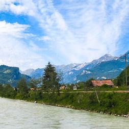 Türkisfarbene Aare fließt durch das Meiringen-Tal mit schneebedeckten Alpengipfeln, grünen bewaldeten Hängen und einem Dorf im Hintergrund.