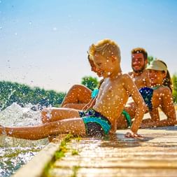 Three children sitting on a wooden dock splashing water with their feet. Water droplets sparkle in the sunlight against a blue sky and green trees.