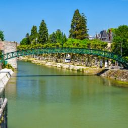 Grüne Metallbrücke Passerelle Victor Hugo über einem Kanal in Montargis, Frankreich, mit Steinmauern, Bäumen und historischen Gebäuden.