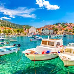 Hvar port with white boats in crystal-clear turquoise water, palm trees, Mediterranean buildings, and green hills under blue sky.