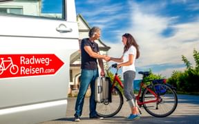 Two women shaking hands next to a bicycle and Radweg-Reisen.com branded van. One woman holds luggage, the other stands with a touring bike.