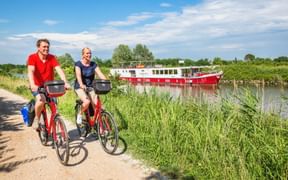 Zwei Radfahrer auf roten Rädern fahren auf einem Schotterweg neben einem Kanal mit einem rot-weißen Flusskreuzfahrtschiff in grüner Landschaft.