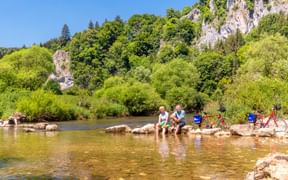 Zwei Radfahrer kühlen ihre Füße in der Donau bei Fridingen, mit roten Tourenrädern am felsigen Ufer und Kalksteinfelsen im Hintergrund.