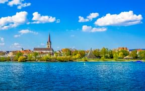Blick über den Bodensee auf Radolfzell mit Kirchturm, historischen Gebäuden und grünen Bäumen entlang des Ufers unter blauem Himmel.