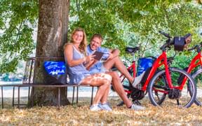 Zwei Radfahrer sitzen auf einer Bank unter einem Baum in Stein am Rhein und schauen auf ein Tablet. Zwei rote E-Bikes mit blauen Taschen stehen daneben.