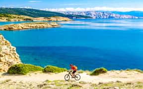Radfahrer in roter Jacke fährt entlang Küstenweg mit Blick auf türkisfarbene Adria mit Felsklippen und Bergen in Kroatien.