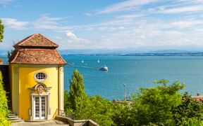 Gelber Barockpavillon mit Ziegeldach im Meersburger Schlossgarten mit Blick auf den Bodensee. Gepflegte Hecken und Topfbäume rahmen die Aussicht.