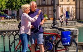 Senior couple with bicycles on a bridge in Interlaken, pointing at sights. Turquoise canal and historic buildings visible in background.